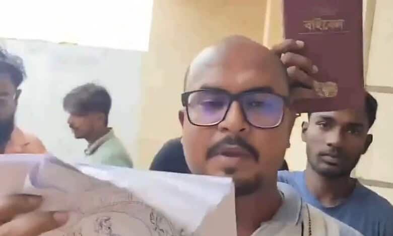 Man holding a book with others in the background, indoors, serious expression.