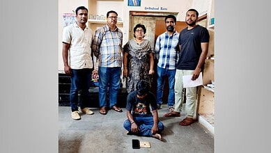 Group of five men and one woman standing in a room, one man sitting on the floor with a mobile phone.