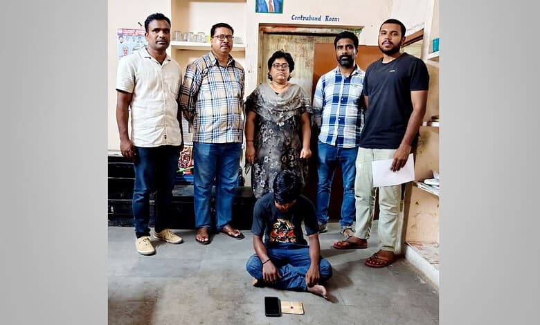 Group of five men and one woman standing in a room, one man sitting on the floor with a mobile phone.