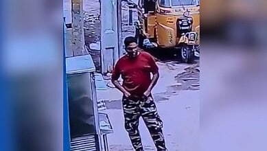 Man in red shirt and camouflage pants on street near yellow auto rickshaw.