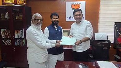 Three men in an office exchanging a document, with a BJP logo in the background.