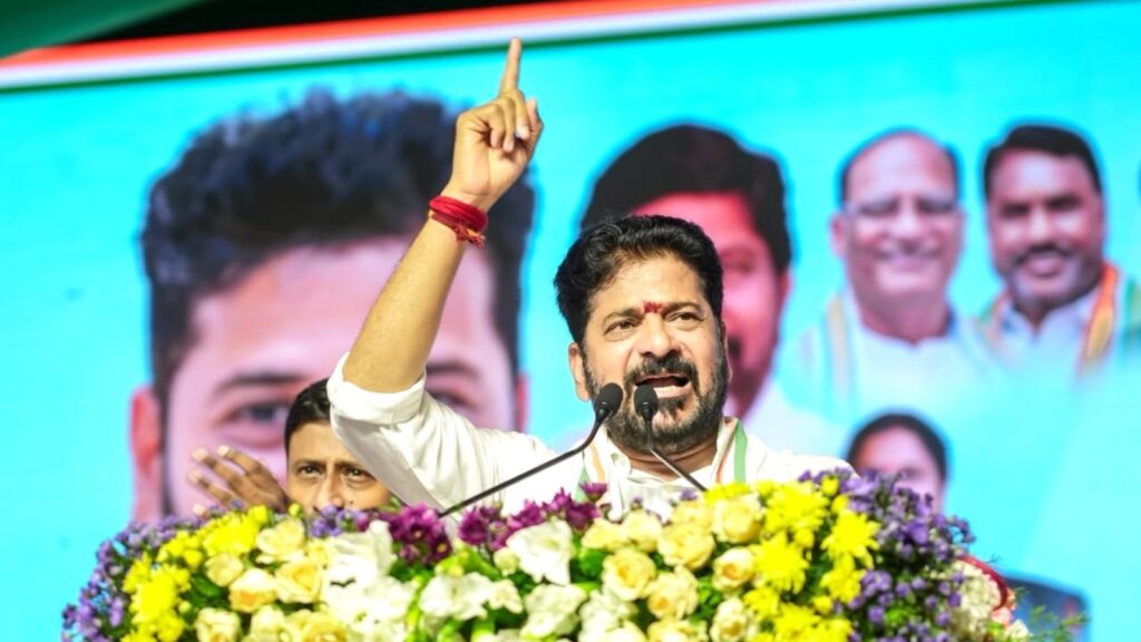 Revanth Reddy speaking at a political rally with a large screen behind him showing his image.
