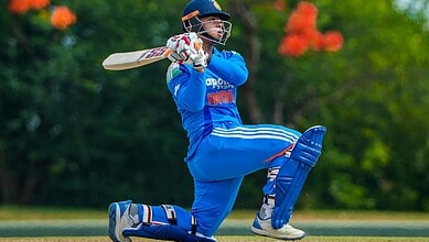 Cricketer in blue uniform playing a shot on the cricket field with trees in background.