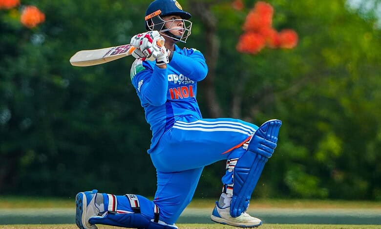 Cricketer in blue uniform playing a shot on the cricket field with trees in background.