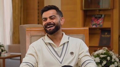 Smiling man with a beard in a cream shirt sitting in a wooden room.