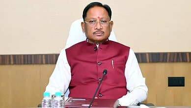A senior Indian official wearing a maroon waistcoat and white shirt, seated at a desk with microphones an.
