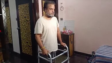 A man using a walker indoors, with a serious expression, in a modest room with a bed and a small table.