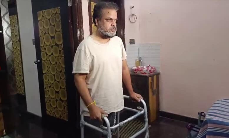 A man using a walker indoors, with a serious expression, in a modest room with a bed and a small table.