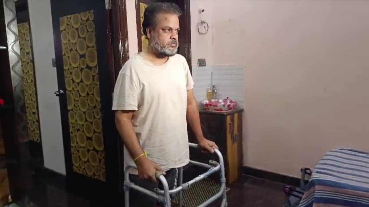 A man using a walker indoors, with a serious expression, in a modest room with a bed and a small table.