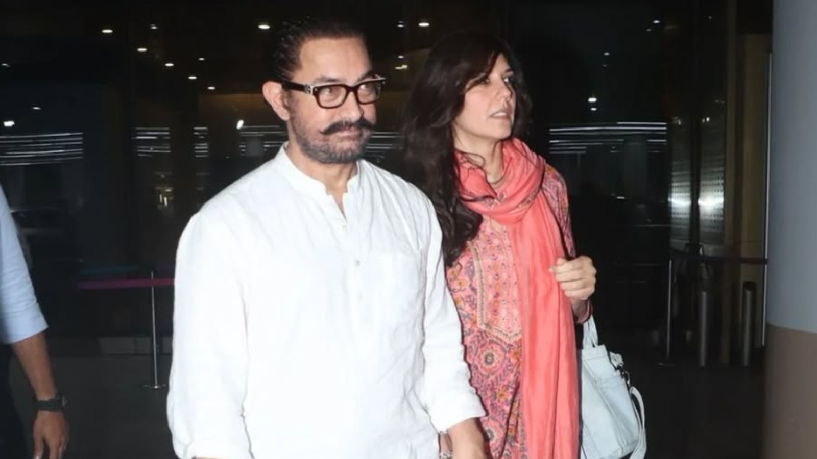 A man with glasses and a woman in a pink scarf walking through an airport terminal.