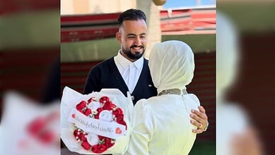 Smiling man in a black sweater hugging a woman in white with a headscarf.