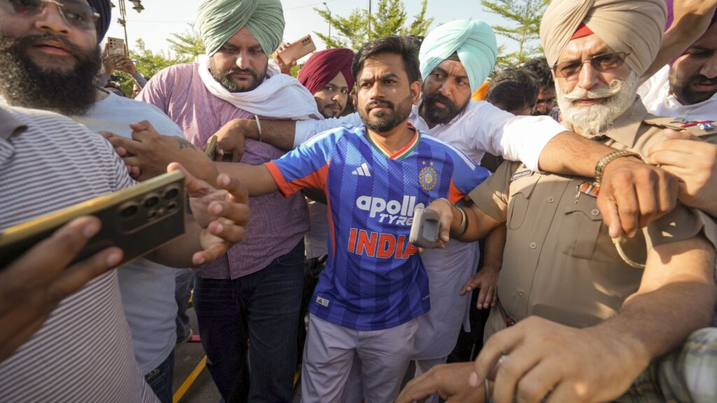 A woman in a blue India cricket jersey surrounded by a crowd of men, some wearing turbans, in an outdoor.