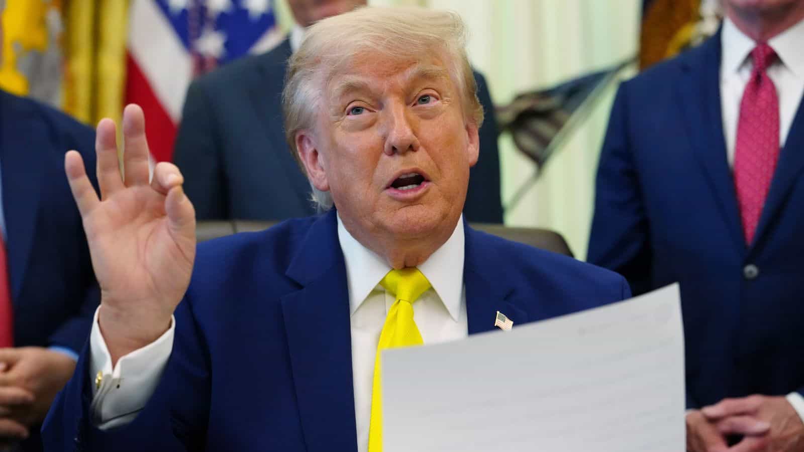 Donald Trump speaking and gesturing with his hand during a press event, surrounded by officials in the Wh.