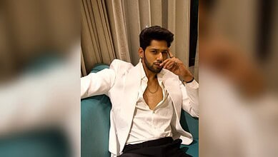 Baseer Ali in a white blazer and shirt, sitting on a blue sofa, looking thoughtful.
