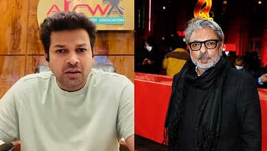 Two men seated at an event, one in a light shirt, the other in black with glasses and grey hair.