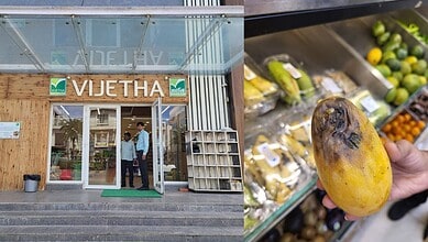 Supermarket entrance with two people and a hand holding a yellow mango near produce shelves.