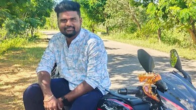 Man sitting on motorcycle with trees and road in background.