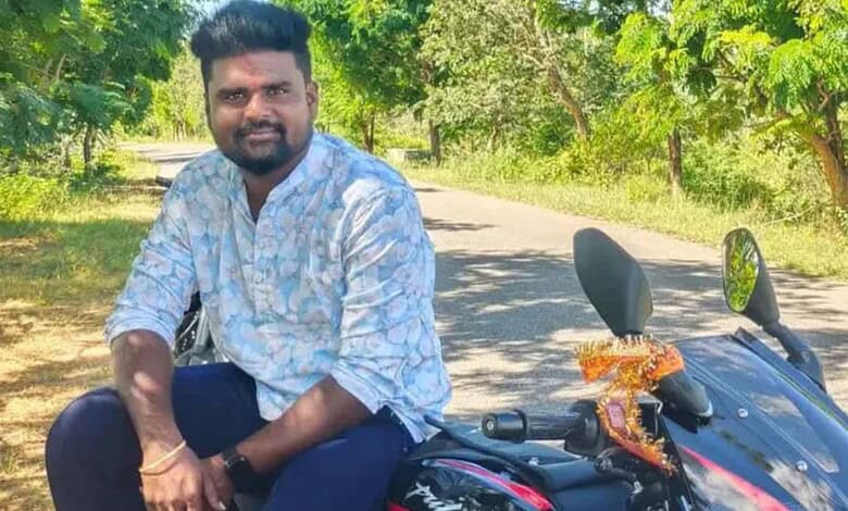Man sitting on motorcycle with trees and road in background.