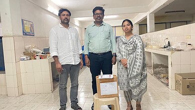 A woman and two men standing in a kitchen or food preparation area, with boxes and supplies around them.