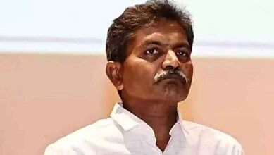 Indian politician with mustache in white shirt during a public event, serious expression.