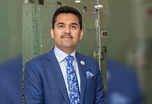 Businessman in a blue suit smiling at a professional event, standing in front of a glass wall.