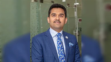 Businessman in a blue suit smiling at a professional event, standing in front of a glass wall.