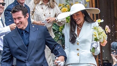 A woman in a white outfit and large hat holding a bouquet, smiling with a man in a blue suit at a joyful.
