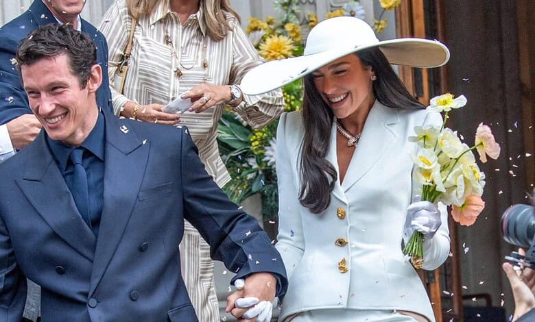 A woman in a white outfit and large hat holding a bouquet, smiling with a man in a blue suit at a joyful.