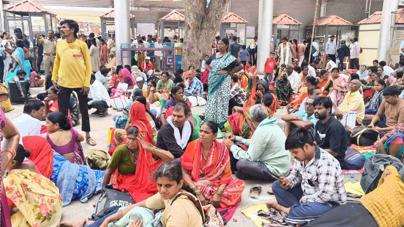 A busy outdoor gathering with diverse people sitting on the ground, some engaging in conversations, under.