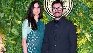 A woman and a man standing outdoors in front of lush green foliage and a decorative circular emblem.