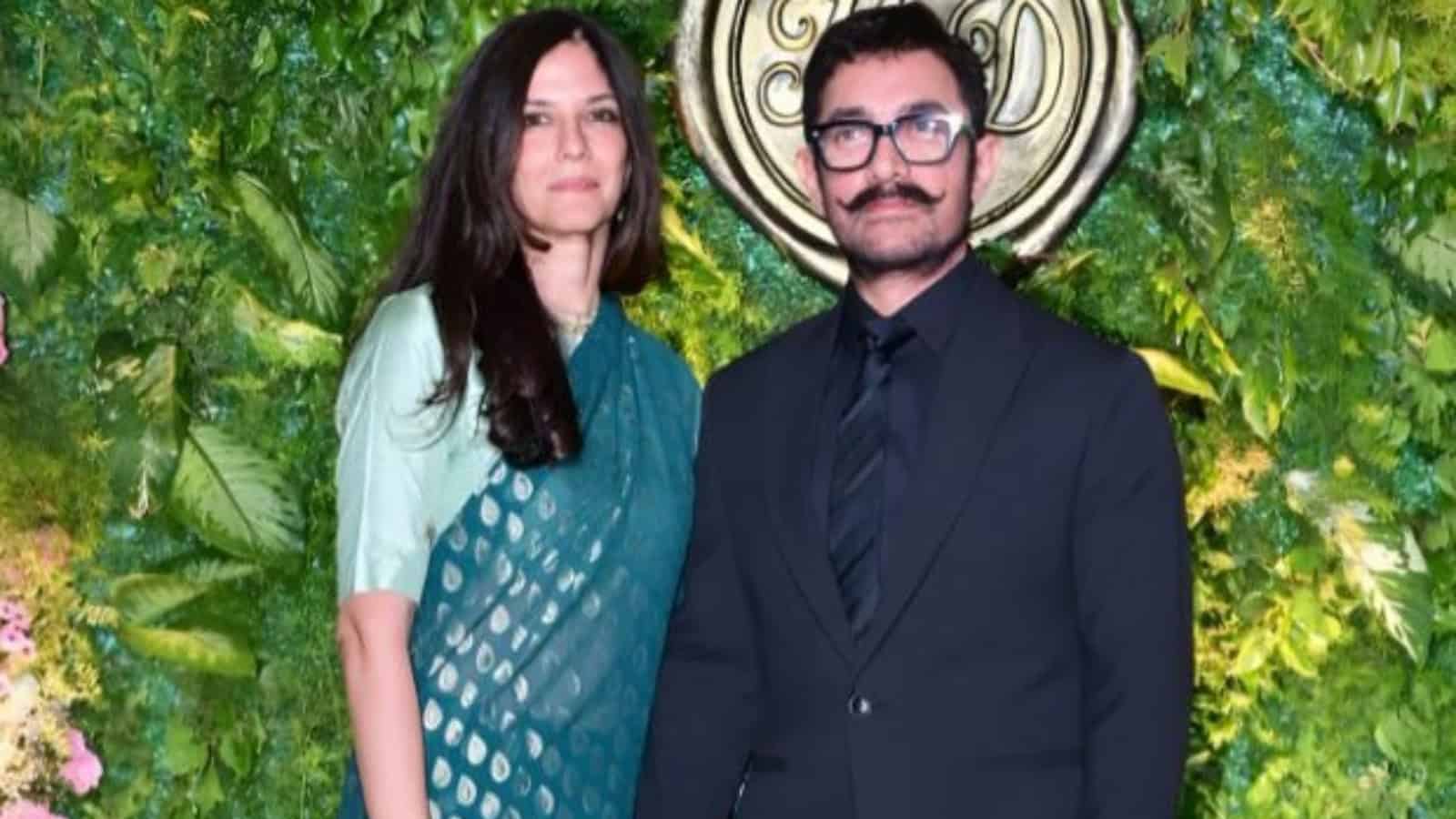 A woman and a man standing outdoors in front of lush green foliage and a decorative circular emblem.
