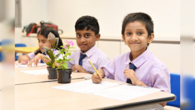 Schoolchildren sit in a classroom, writing at their desks beside potted plants during a learning activity.