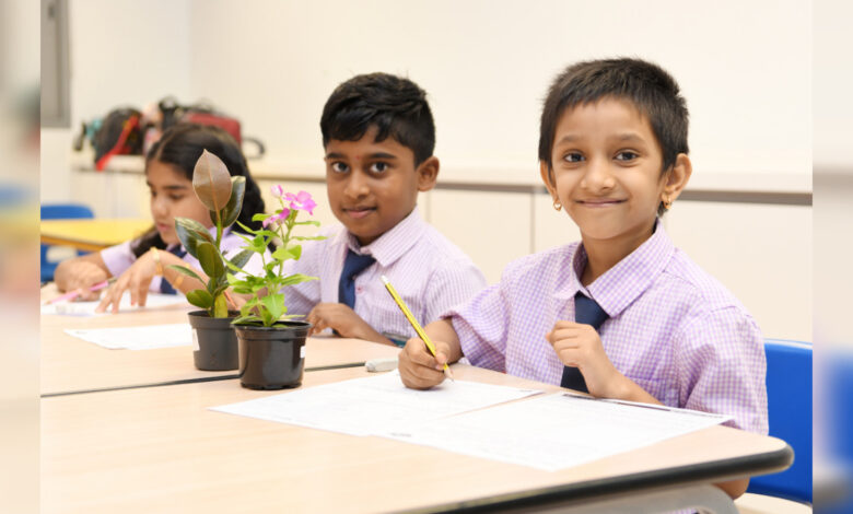 Schoolchildren sit in a classroom, writing at their desks beside potted plants during a learning activity.