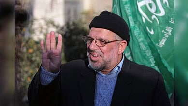 A man wearing glasses and a black cap waving, with a green flag in the background, participating in a pub.