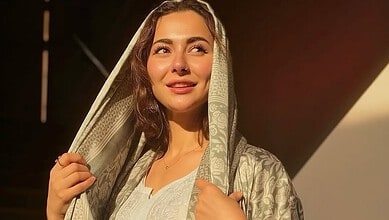 A woman with long brown hair wearing a traditional light-colored shawl, smiling softly in warm sunlight.