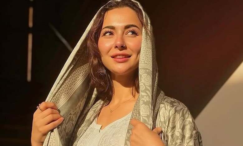 A woman with long brown hair wearing a traditional light-colored shawl, smiling softly in warm sunlight.