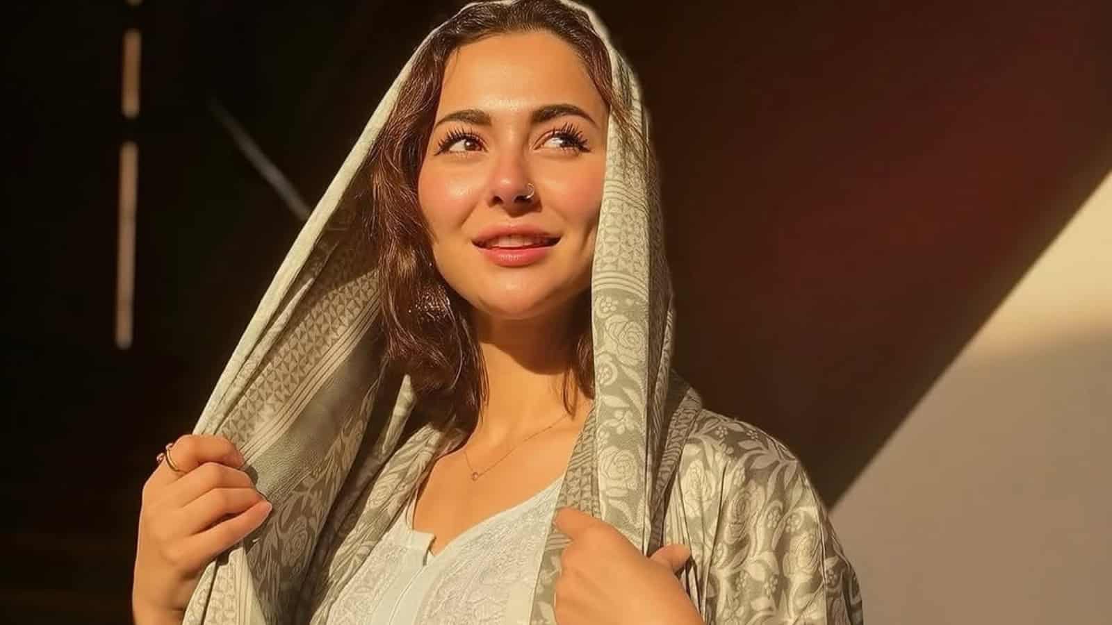 A woman with long brown hair wearing a traditional light-colored shawl, smiling softly in warm sunlight.