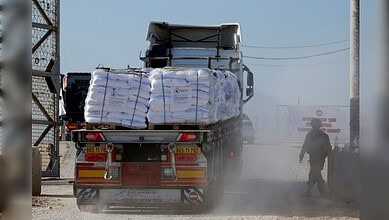 Heavy truck carrying cargo on dusty road with worker nearby in an industrial setting.