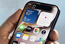 Smartphone displaying weather, calendar, and app icons, held in a person's hand against a blurred outdoor.