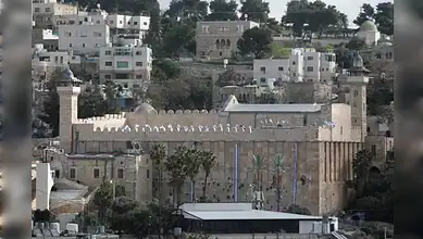 Ancient city wall and gate in Hebron, Palestine, with surrounding buildings and greenery.