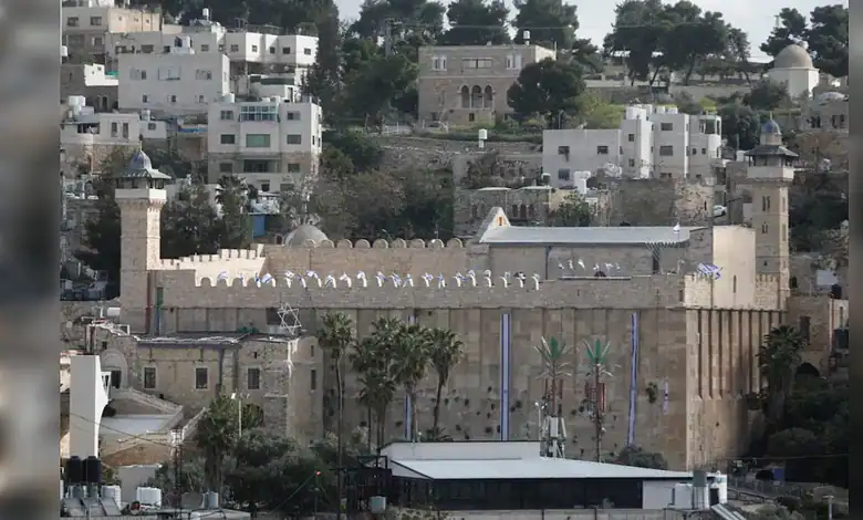 Ancient city wall and gate in Hebron, Palestine, with surrounding buildings and greenery.