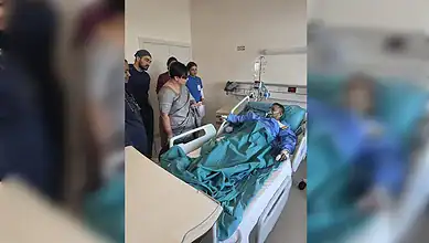 Patient in hospital bed with medical staff and visitors in a healthcare setting.