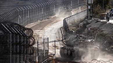 Tank moving on a dusty construction site with fencing and workers in the background.