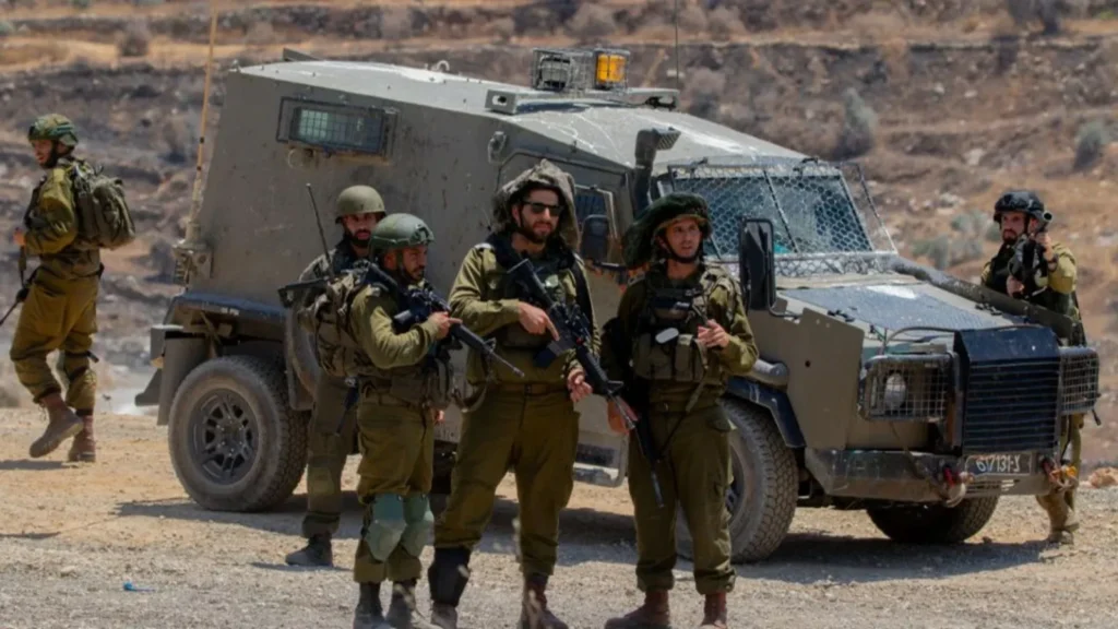 Group of soldiers in military gear standing beside an armored vehicle in a desert landscape.