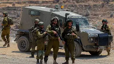 Group of soldiers in military gear standing beside an armored vehicle in a desert landscape.