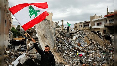 Man holding Lebanese flag in front of rubble and damaged buildings.