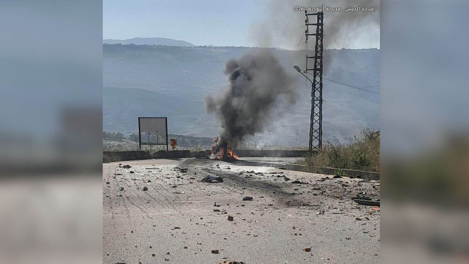 Fire and smoke emerging from a vehicle or object on the road, with electrical infrastructure in the backg.