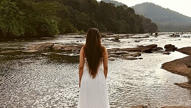A woman with long hair wearing a white dress standing by a river with rocks and lush green trees in the b.
