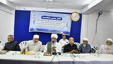 Group of men at a memorial event, some wearing traditional Islamic attire, seated at a table with a blue/.
