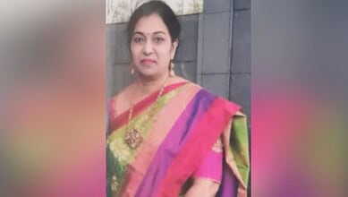 A woman in a colourful saree standing outdoors against a grey wall.